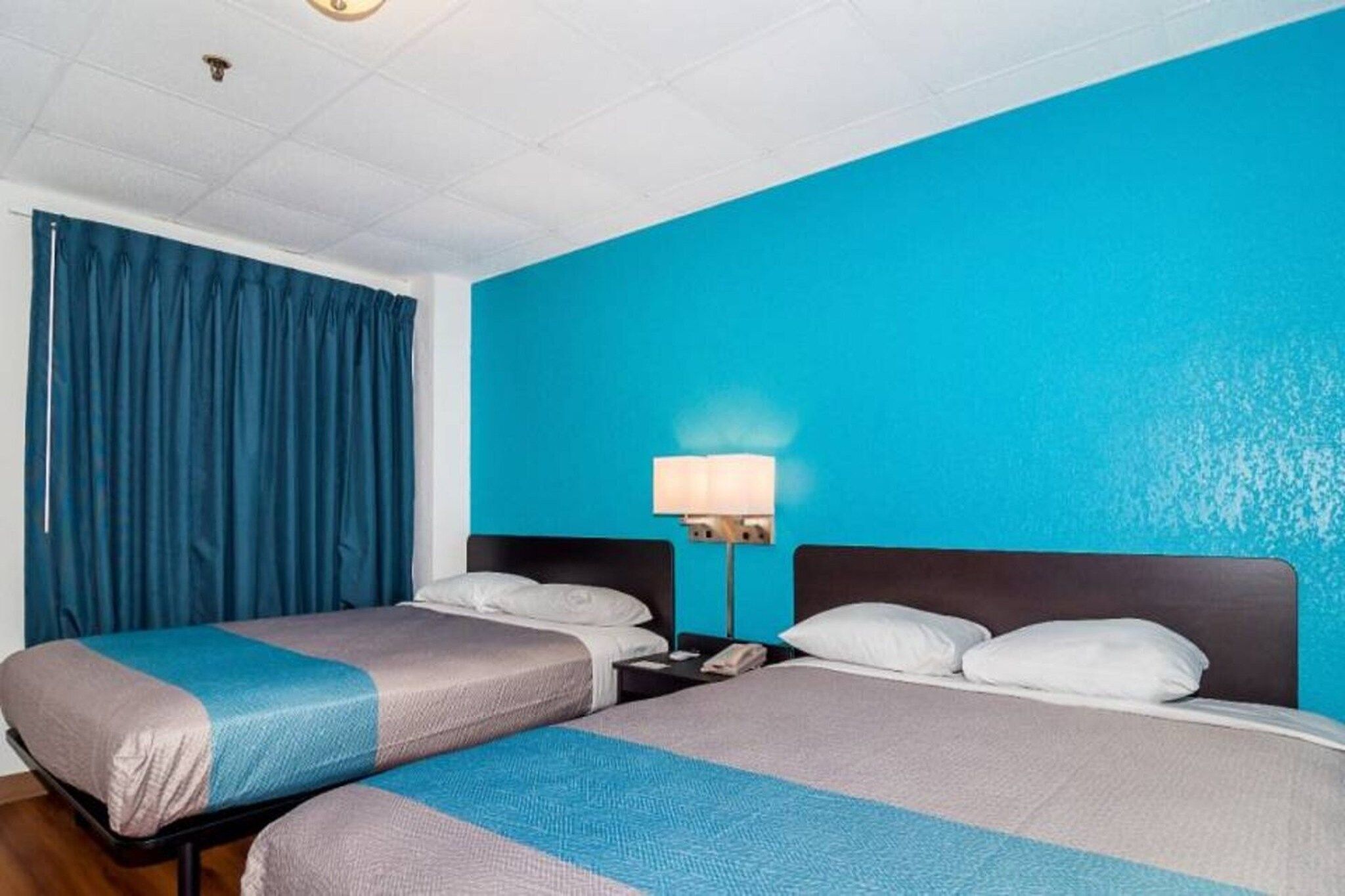 Quarto de hotel com duas camas, uma mesa de cabeceira, uma luminária e cortina azul.