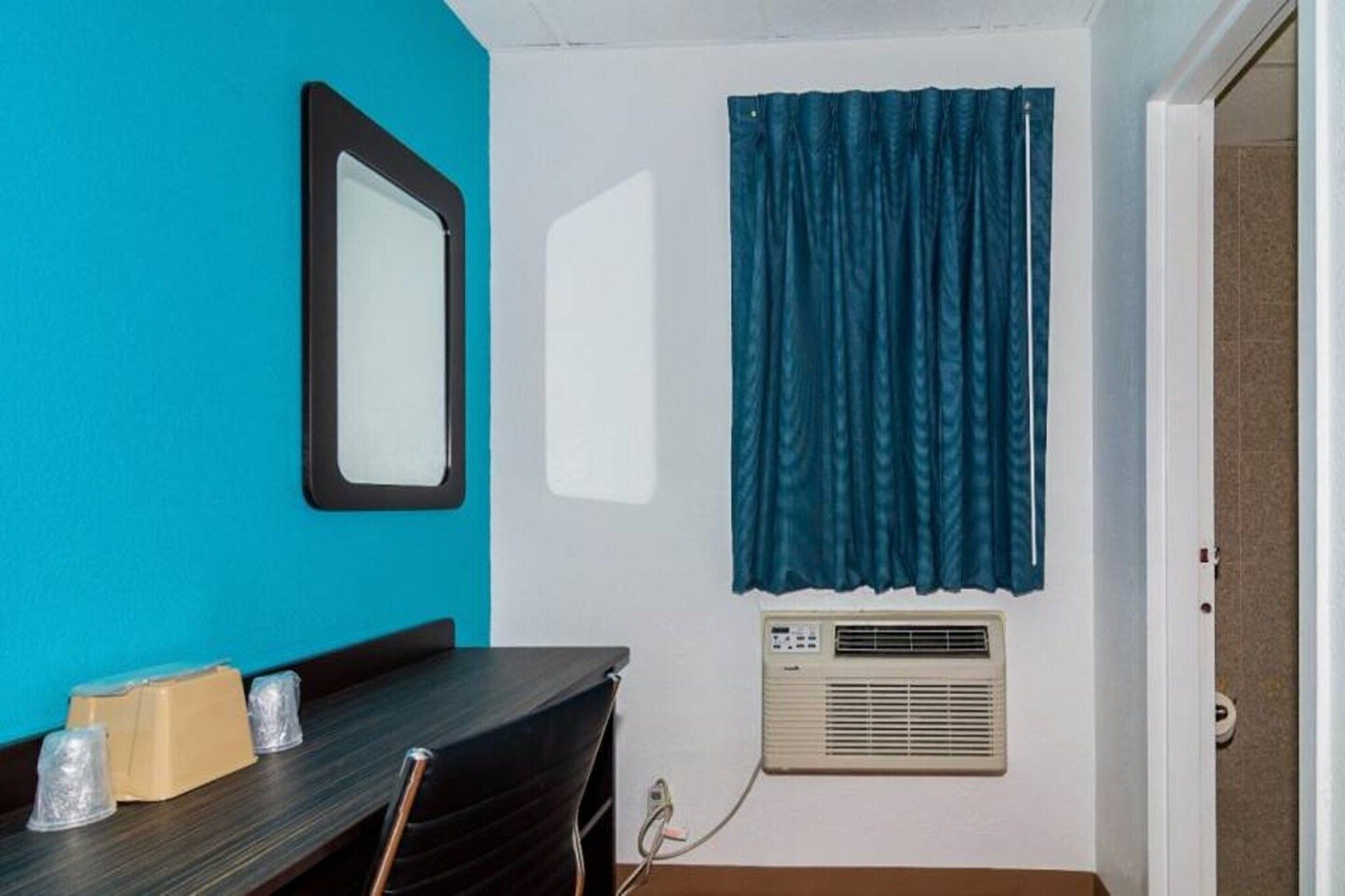 A room with a teal wall, a black-framed mirror, a window with blue curtains, a table with chairs, and a wall-mounted air conditioner.