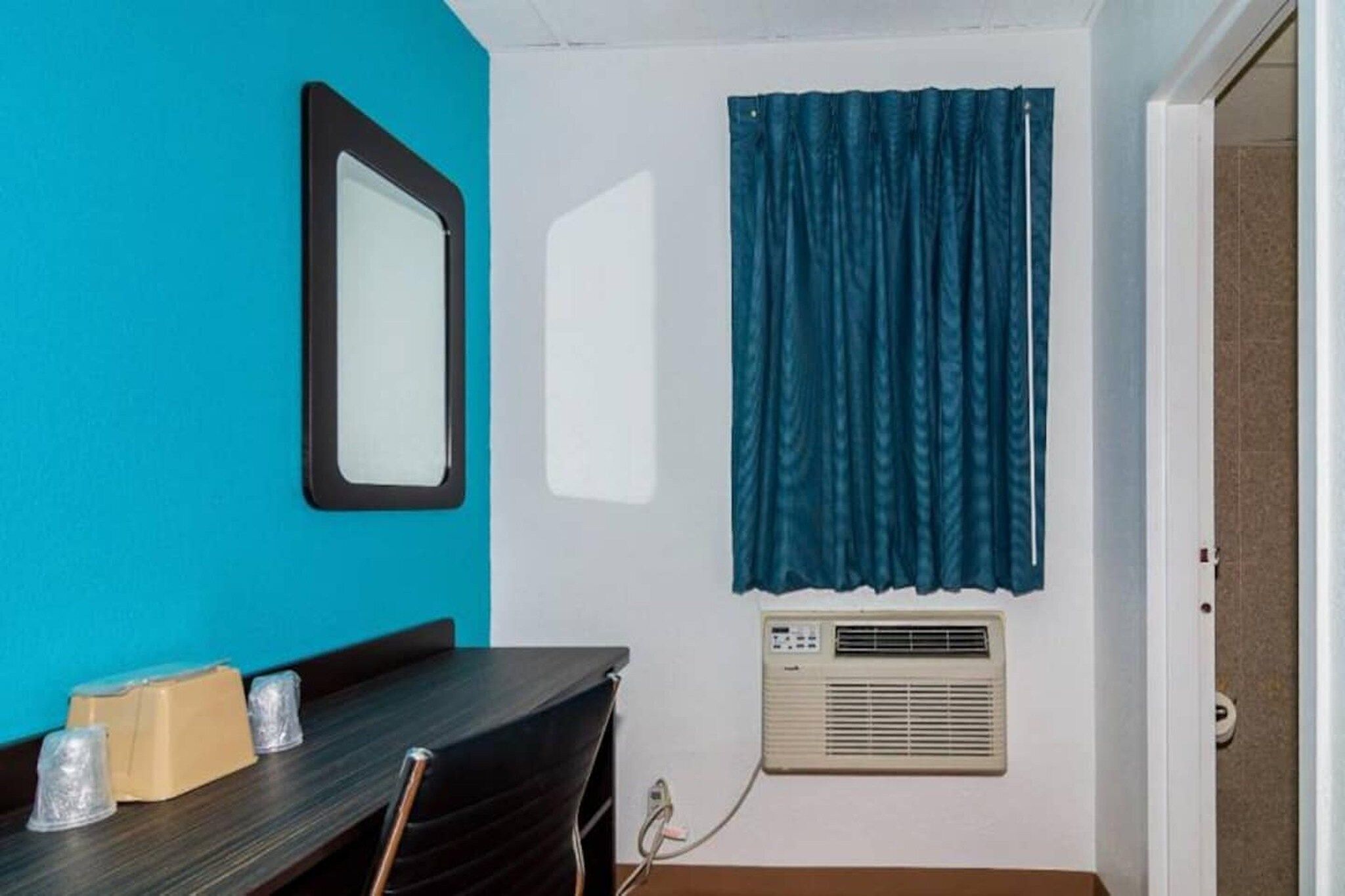 Ein Zimmer mit Tisch, Stühlen, einem Spiegel, einem Fenster mit Vorhängen und einer Klimaanlage.