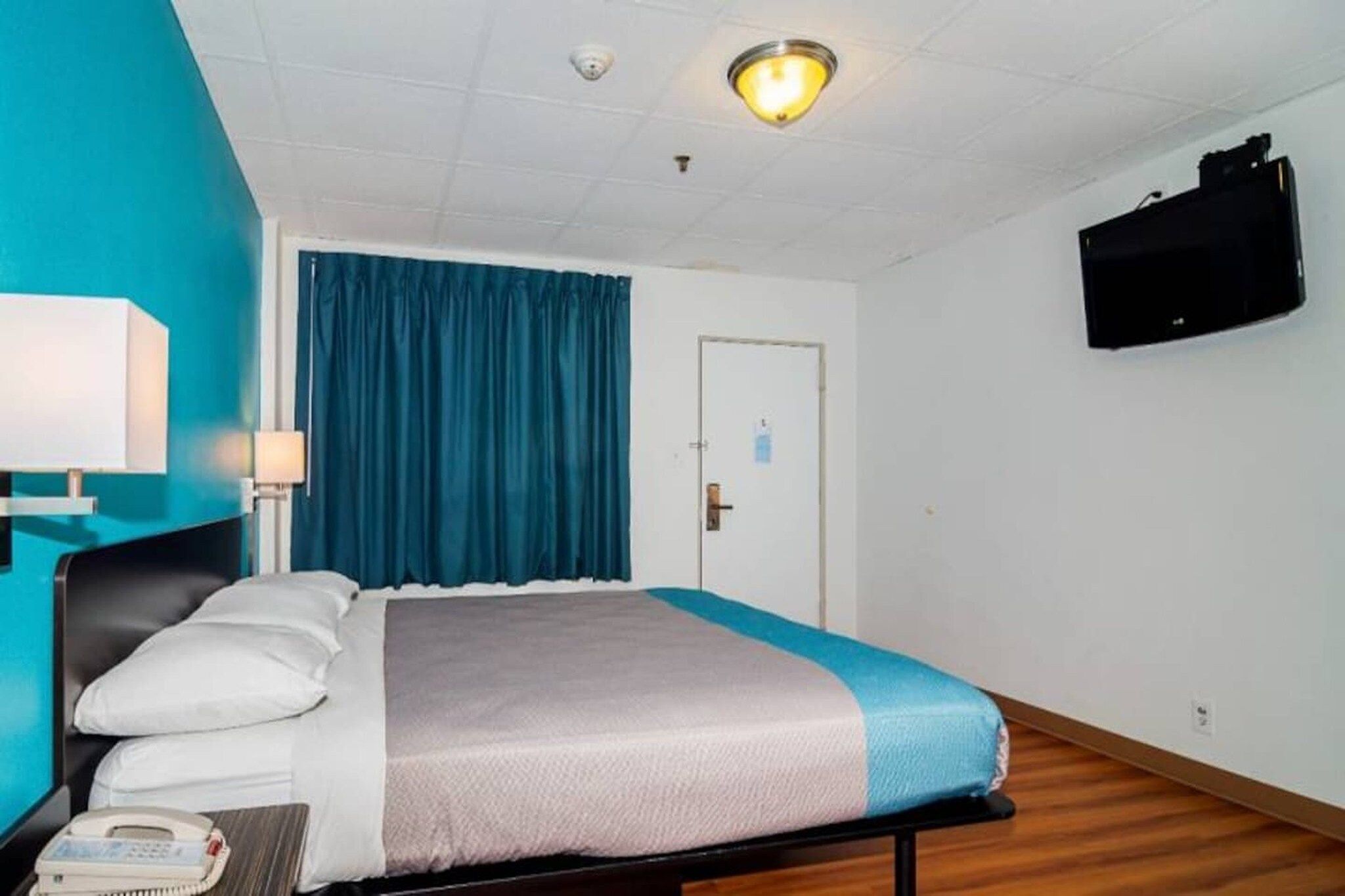 A hotel room with a bed, a desk with a telephone, a wall-mounted TV, and a door with a lock.