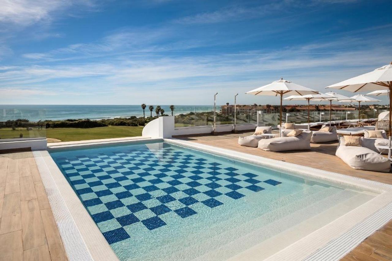 Una piscina con diseño de cuadros, rodeada de sillones de descanso y sombrillas, con vista al océano.