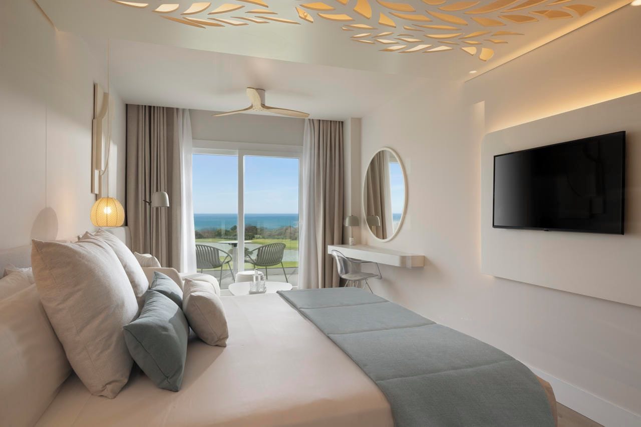 A modern hotel room with a bed, a TV, a balcony with a view, and a ceiling with a decorative pattern.