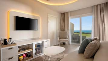 Habitación de hotel moderna con una televisión de pantalla plana, una consola blanca con cajones, una mesita blanca pequeña, un sofá cómodo, un sillón de mimbre y un amplio ventanal con vistas al océano.