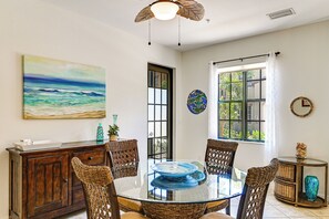 A dining area with a glass-top table surrounded by wicker chairs, a wooden cabinet, and a painting of a beach scene. - Olé at Lely Resort, More Than a Vacation (Naples)