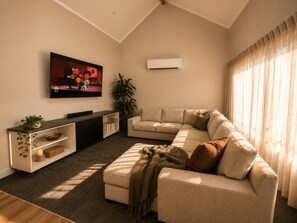 Ein Wohnzimmer mit einer Eckcouch, einem flachbildfernseher an der Wand, einer Topfpflanze und einem großen Fenster mit Vorhängen.