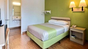 Una habitación de hotel con cama, mesita de noche, lámpara y una pequeña estantería con un teléfono y libros.