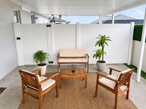 A covered patio with a white fence, a beige sofa, two wooden chairs, a small table, and two potted plants. - Peaceful studio with AC in charming Cape Coral & seasonal pool and jacuzzi (Cape Coral)