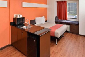 A compact hotel room with a kitchenette, a bed, a window with curtains, and a bench by the window. - 1 King Bed | Smoking (RICHBURG)