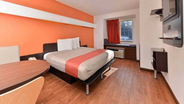 A hotel room with a bed, a desk, a chair, a window with curtains, and a flat-screen TV mounted on the wall. - 1 King Bed | Smoking (RICHBURG)