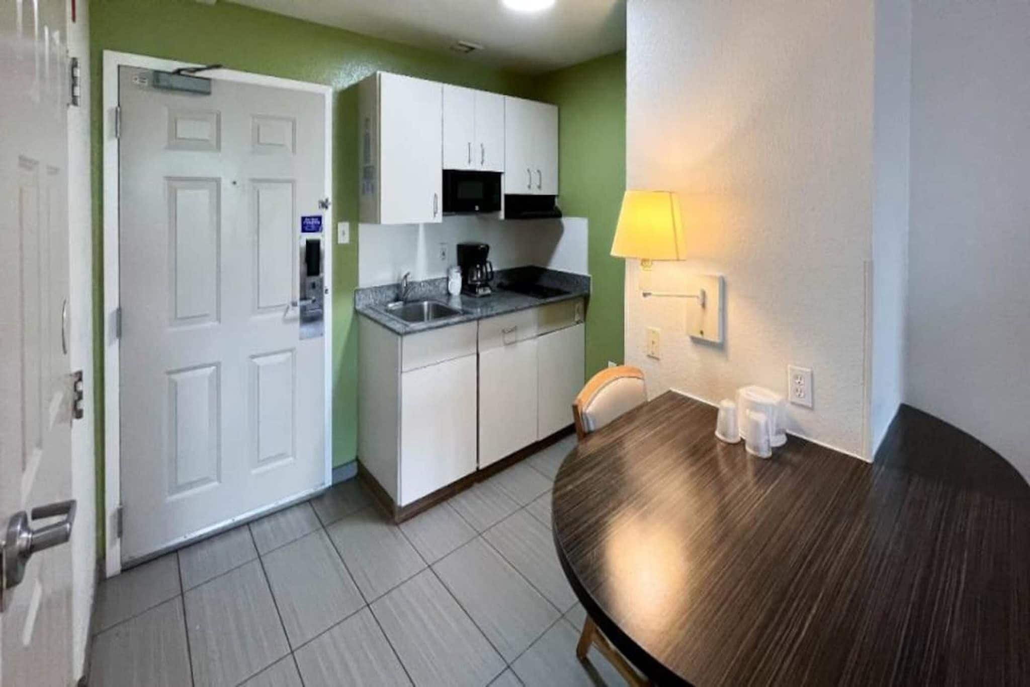 A compact kitchen with white cabinets, a sink, and a coffee maker. There is a dining table with a lamp and cups.