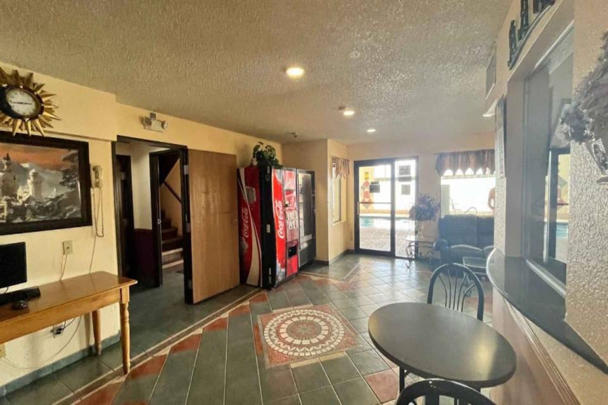 Un vestíbulo de hotel con una recepción, una zona de estar con mesa y sillas, un refrigerador Coca-Cola y un gran ventanal con vistas al área de la piscina.