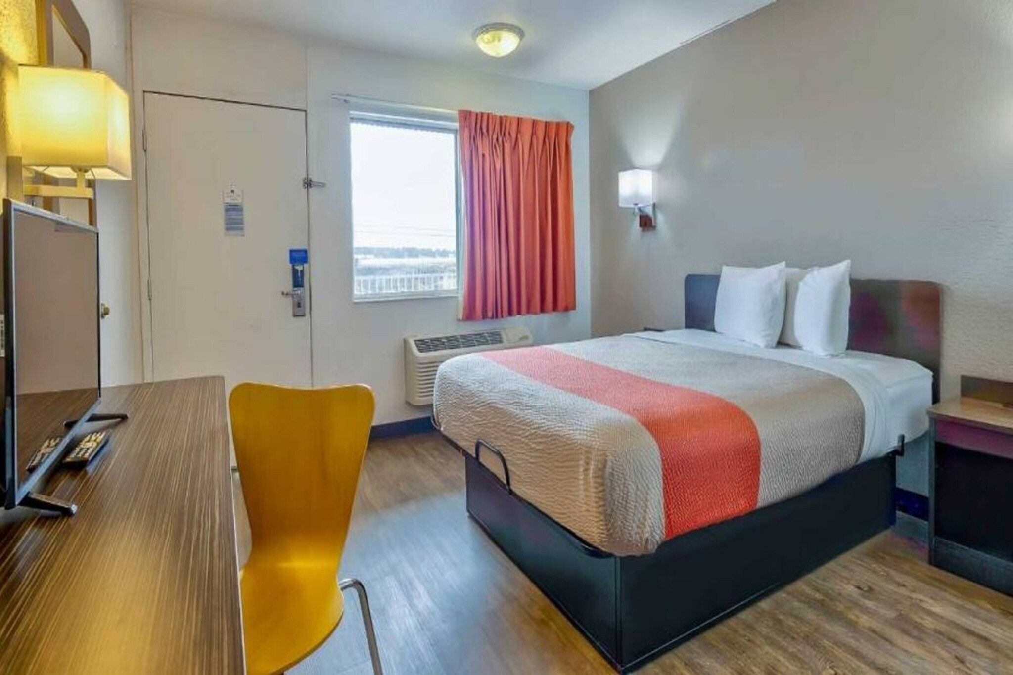 A hotel room with a bed, a desk, a chair, a window with curtains, and a wall-mounted light.