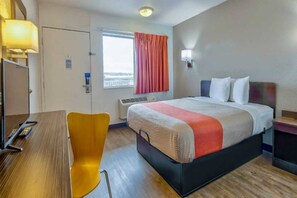A hotel room with a bed, a desk, a chair, a window with curtains, and a wall-mounted light.