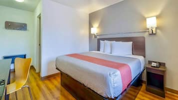 A hotel room with a bed, bedside table, lamp, and a desk with a chair.
