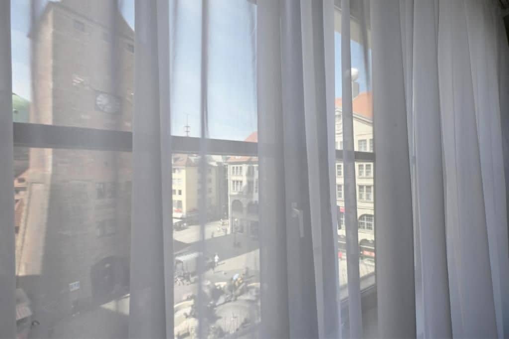 A view through a window with curtains, featuring a street scene with buildings and a clock tower.