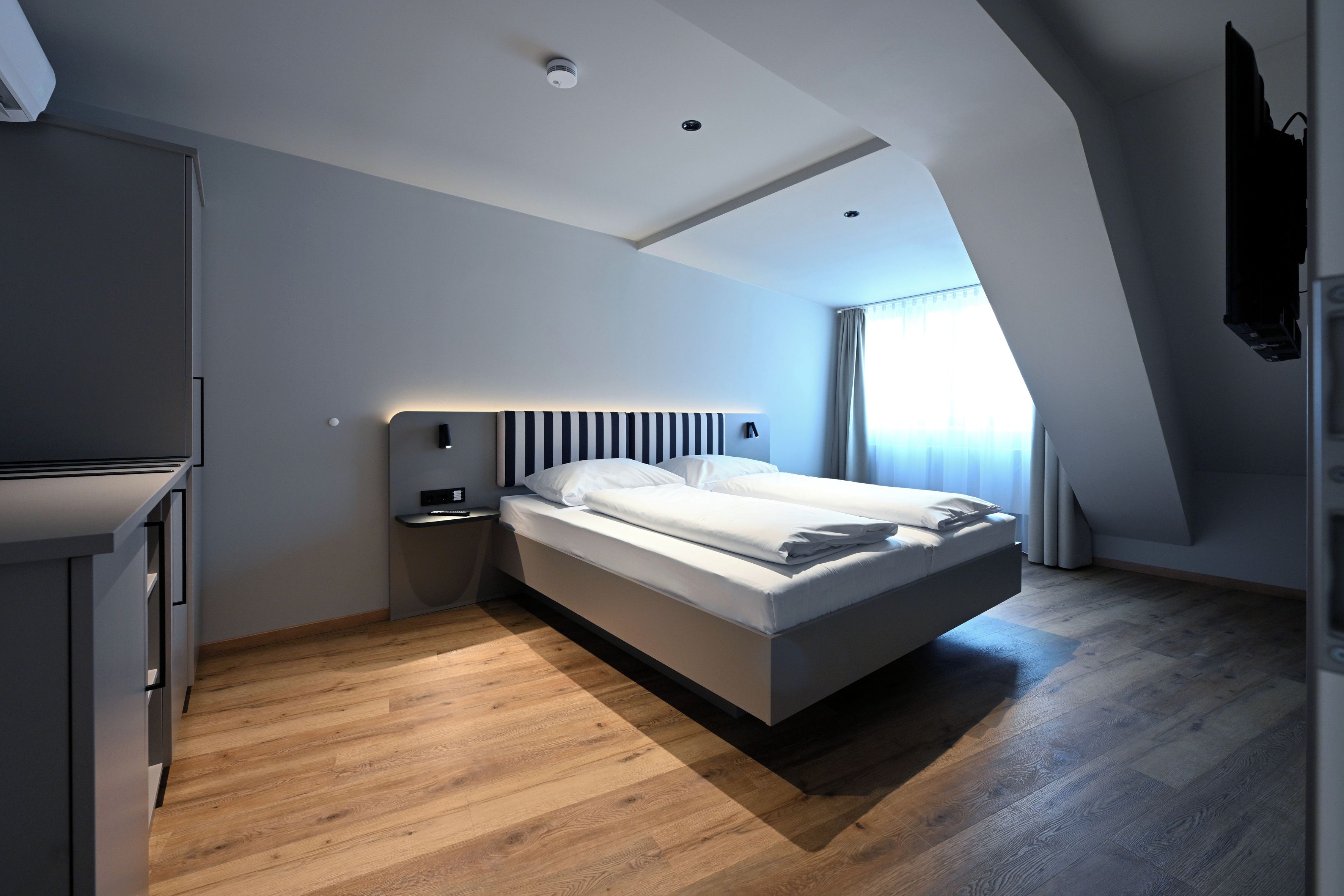 Un dormitorio moderno con techo inclinado, una cama con ropa de cama blanca, una mesita de noche con lámpara y un armario.