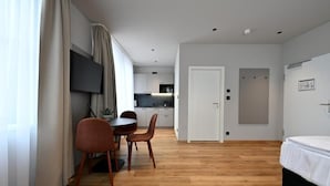 Moderni hotellihuone, jossa on puulattia, sänky, ruokailutila pöytineen ja tuoleineen, seinälle asennettu televisio ja taaempana keittiö.