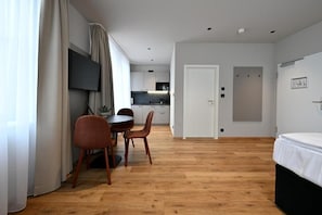 Ein modernes Hotelzimmer mit Holzboden, einem Bett, einem Essbereich mit Tisch und Stühlen, einem an der Wand montierten Fernseher und einer Küche im Hintergrund.