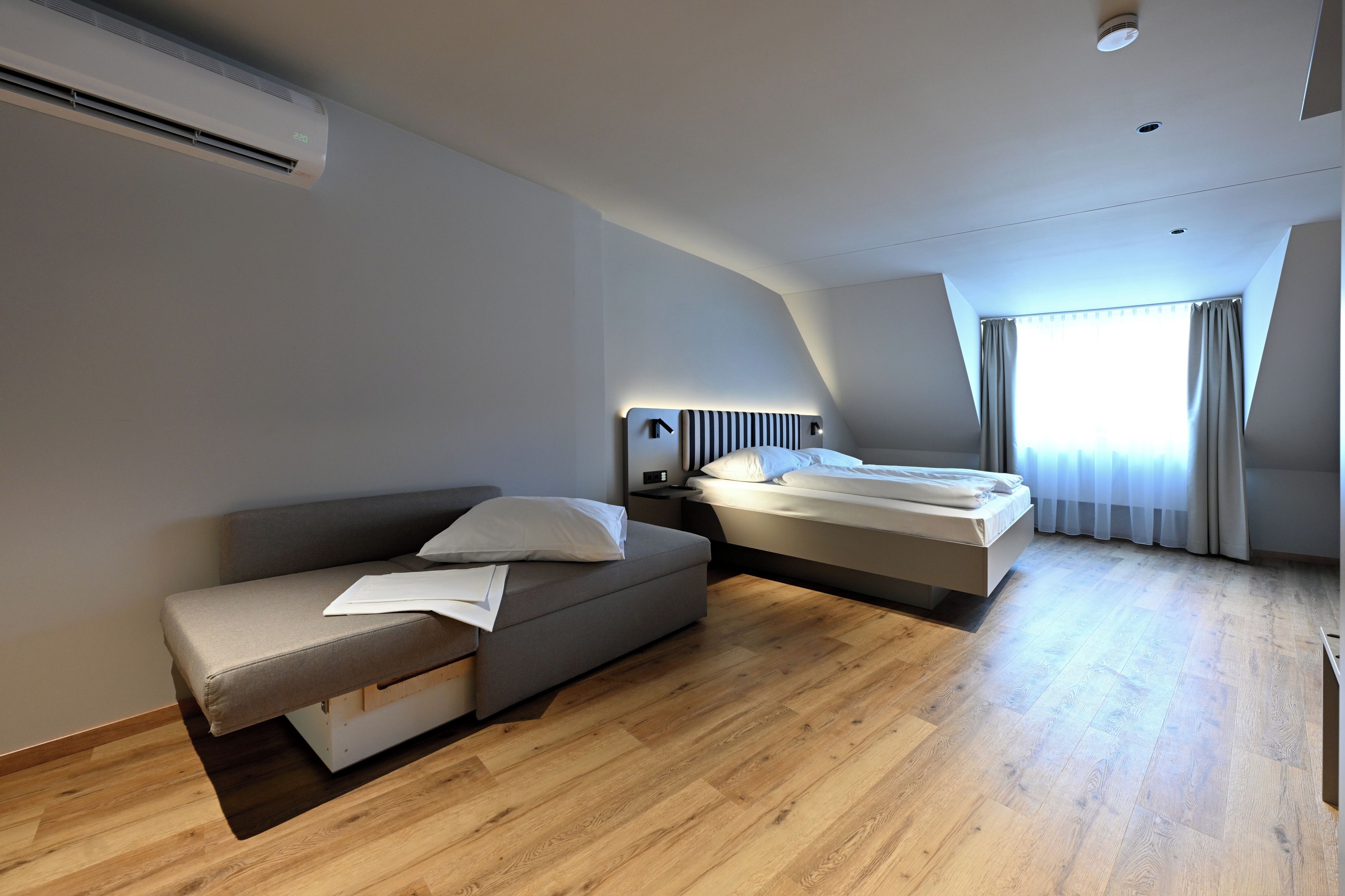 A modern hotel room with a sofa, a bed, a bedside table, and a window with curtains.