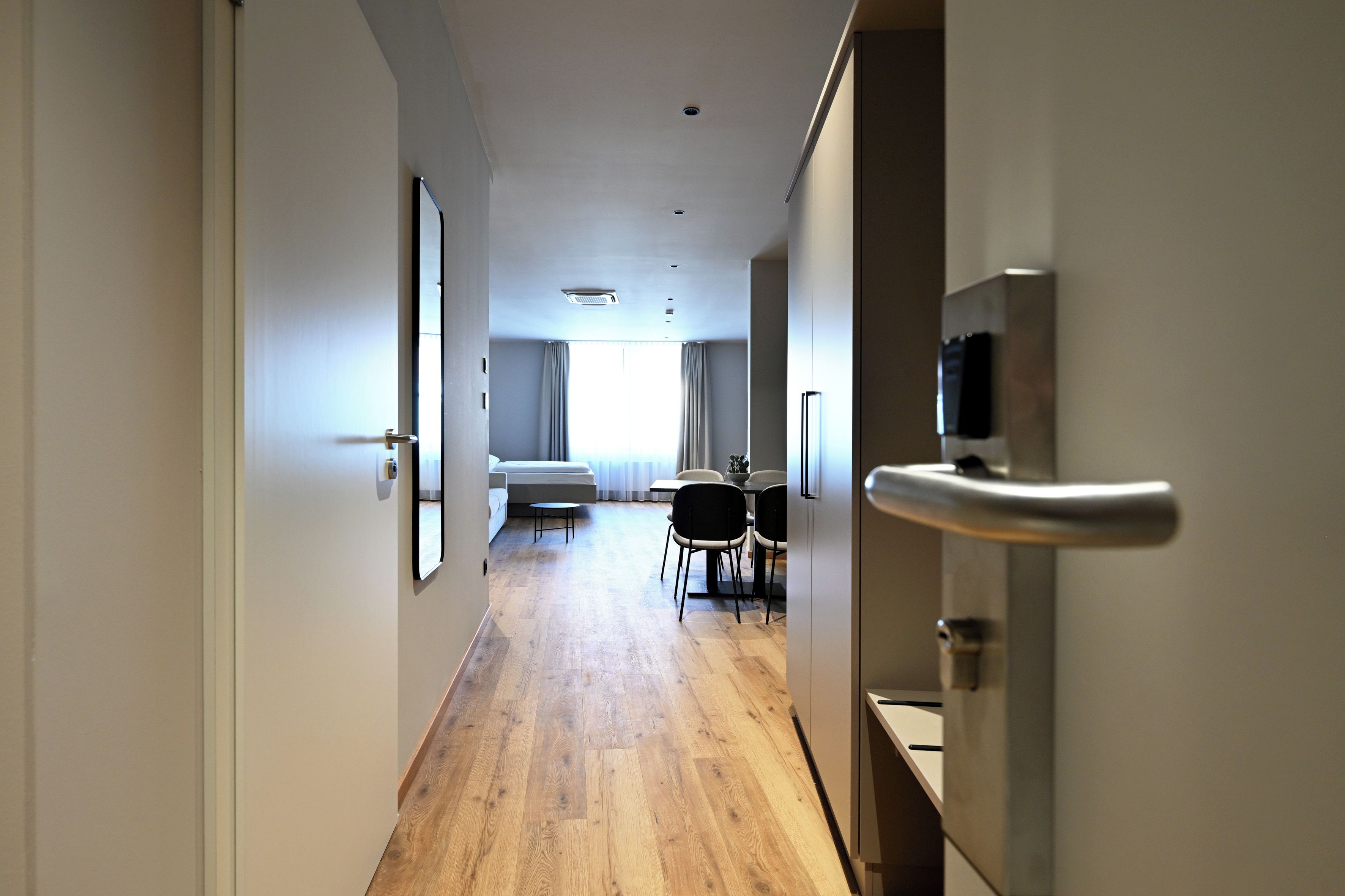 A hotel corridor with a wooden floor, a bed, a dining table with chairs, and a TV mounted on the wall.
