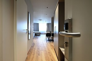 A hotel corridor with a wooden floor, a bed, a dining table with chairs, and a TV mounted on the wall.