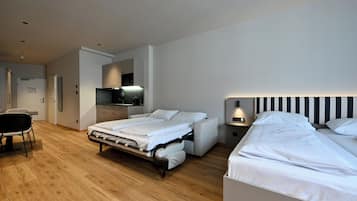 A modern hotel room with a convertible bed, a separate desk area, and a kitchenette.