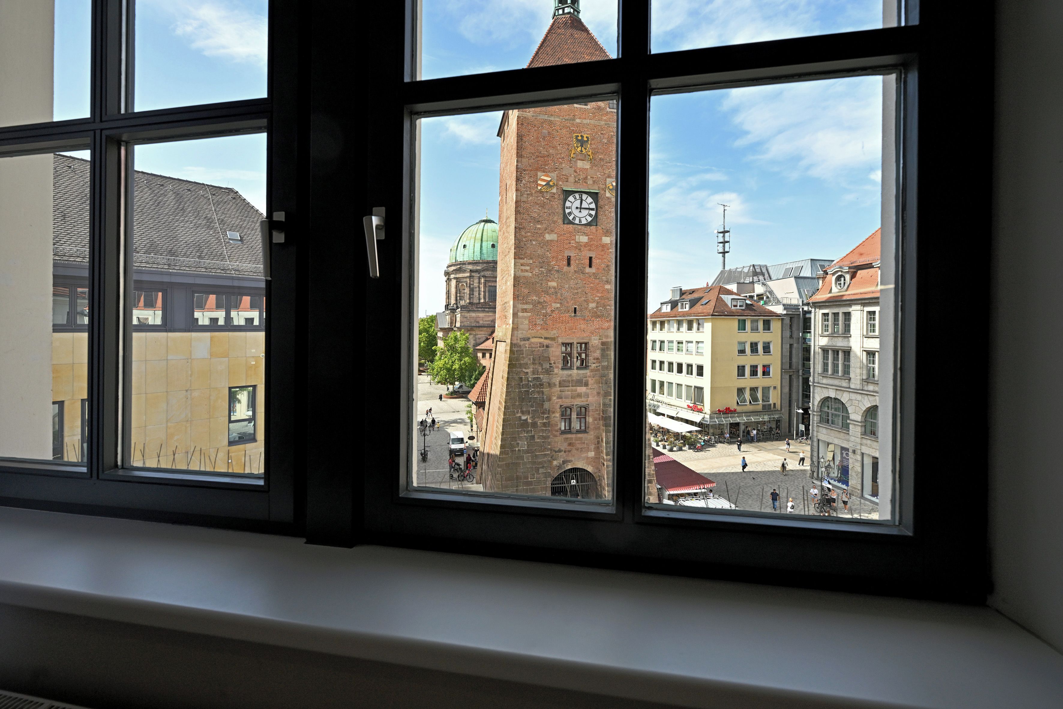 Une fenêtre donnant sur un bâtiment historique doté d’une tour horloge, une rue animée par des piétons et d’autres immeubles.