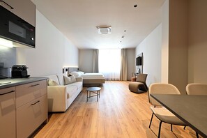 A modern hotel room with a kitchenette, a sofa, a bed, a small table, and a TV.