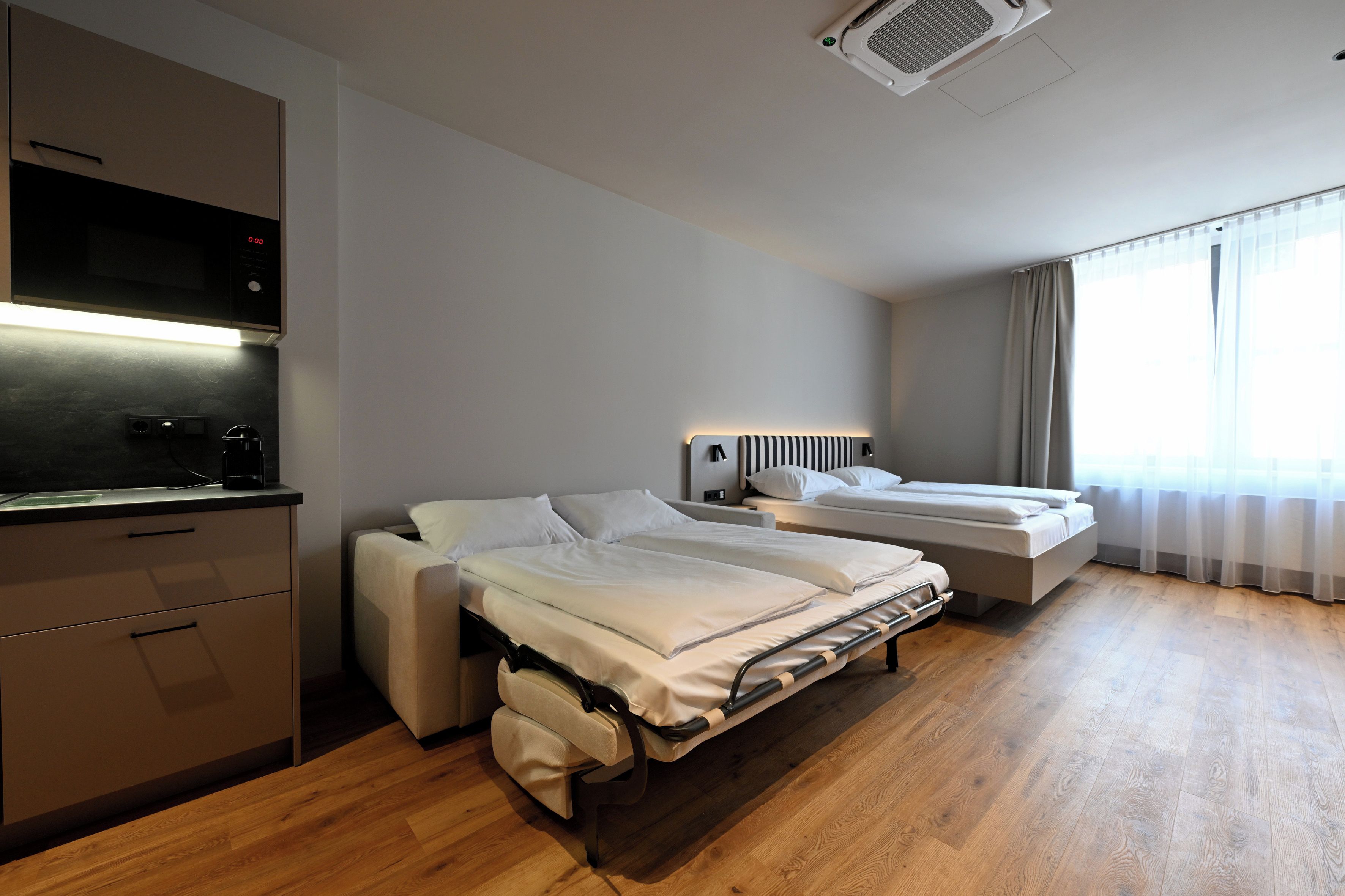 A hotel room with two beds, a kitchenette, and a large window with curtains.
