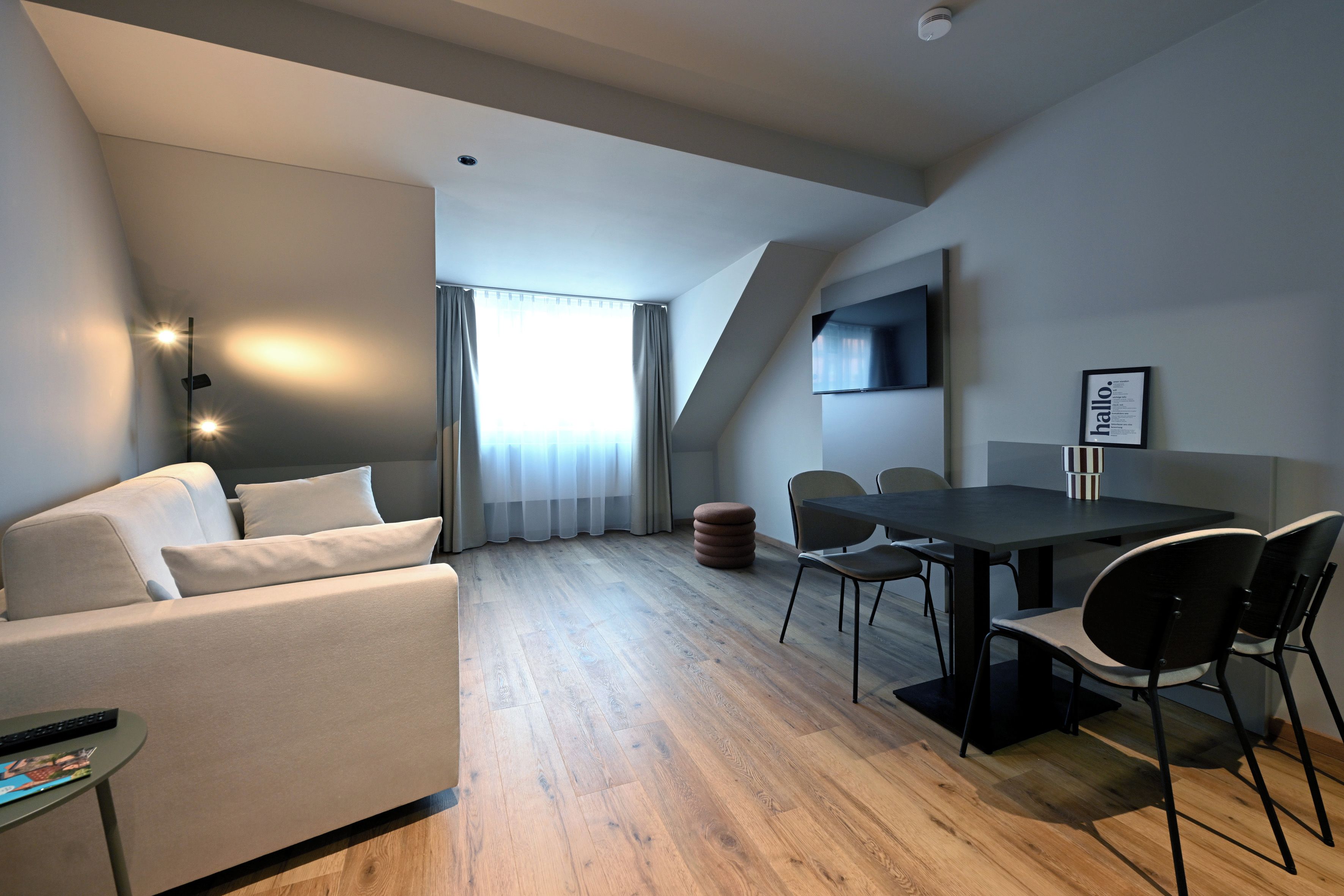 Une pièce moderne et bien éclairée, avec un canapé, une table à manger et des chaises, un téléviseur fixé au mur et une fenêtre avec des rideaux.