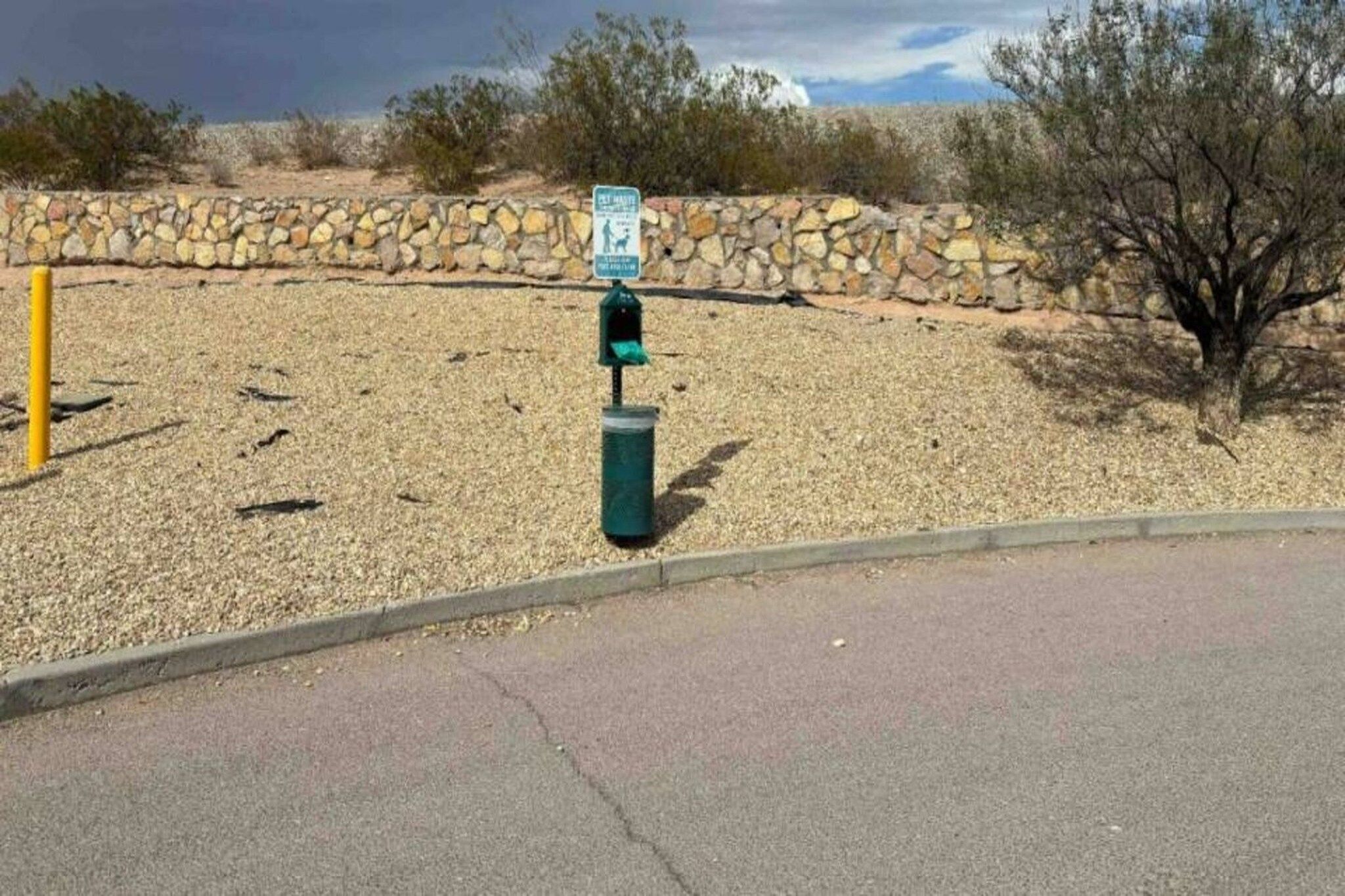 A roadside with a stone wall, a trash can, and a sign.