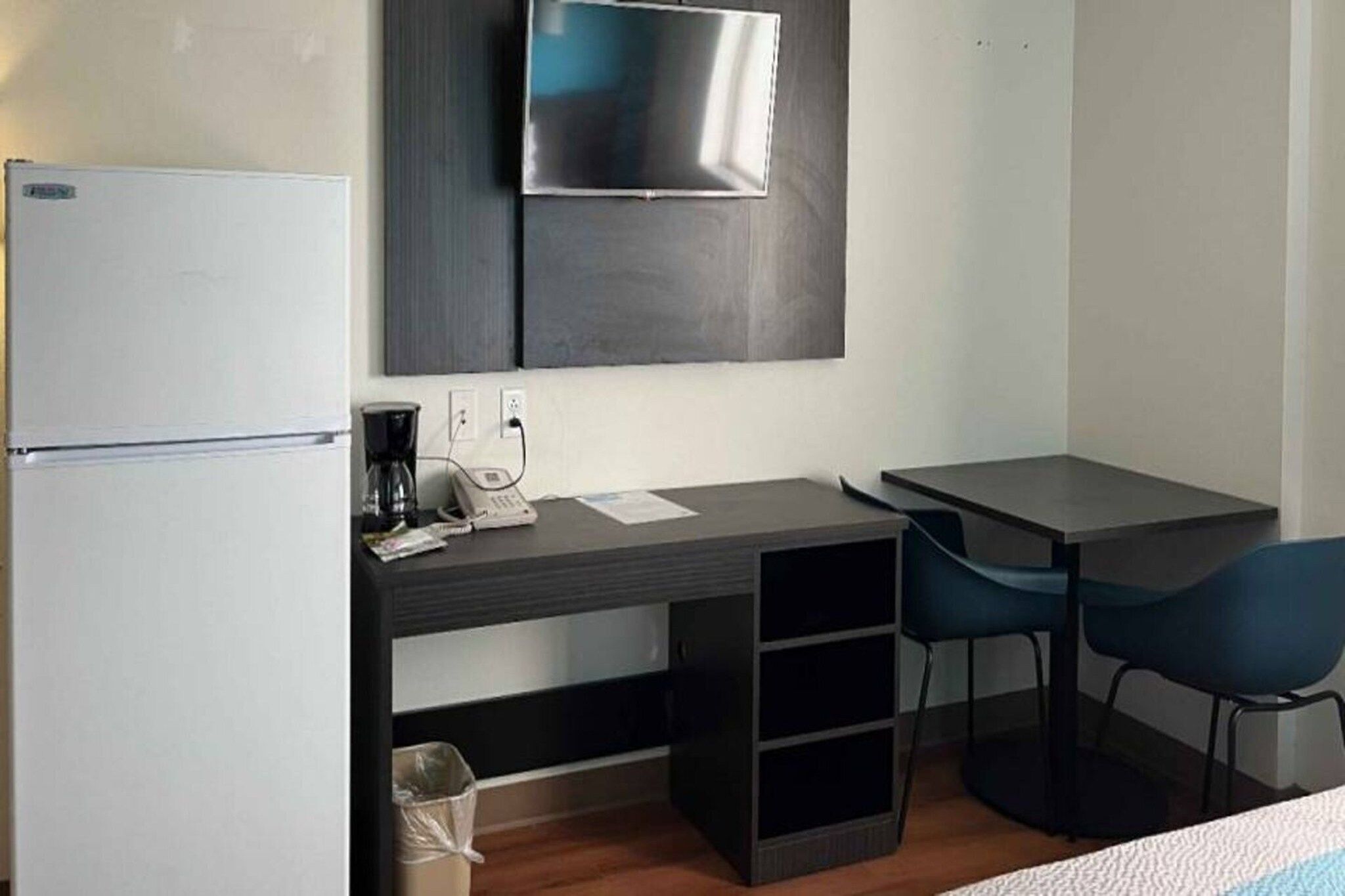 A compact hotel room with a kitchenette, a flat-screen TV mounted on the wall, a desk with a chair, and a small trash bin.