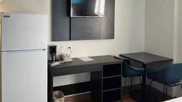 A compact hotel room with a kitchenette, a flat-screen TV mounted on the wall, a desk with a chair, and a small trash bin.