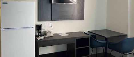 A compact hotel room with a kitchenette, a flat-screen TV mounted on the wall, a desk with a chair, and a small trash bin.