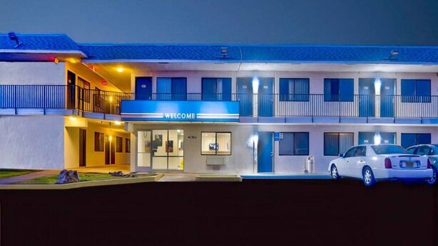 Ein mehrstöckiges Motel mit einem 'WELCOME'-Schild, einem überdachten Parkplatz und einem weißen Auto davor.