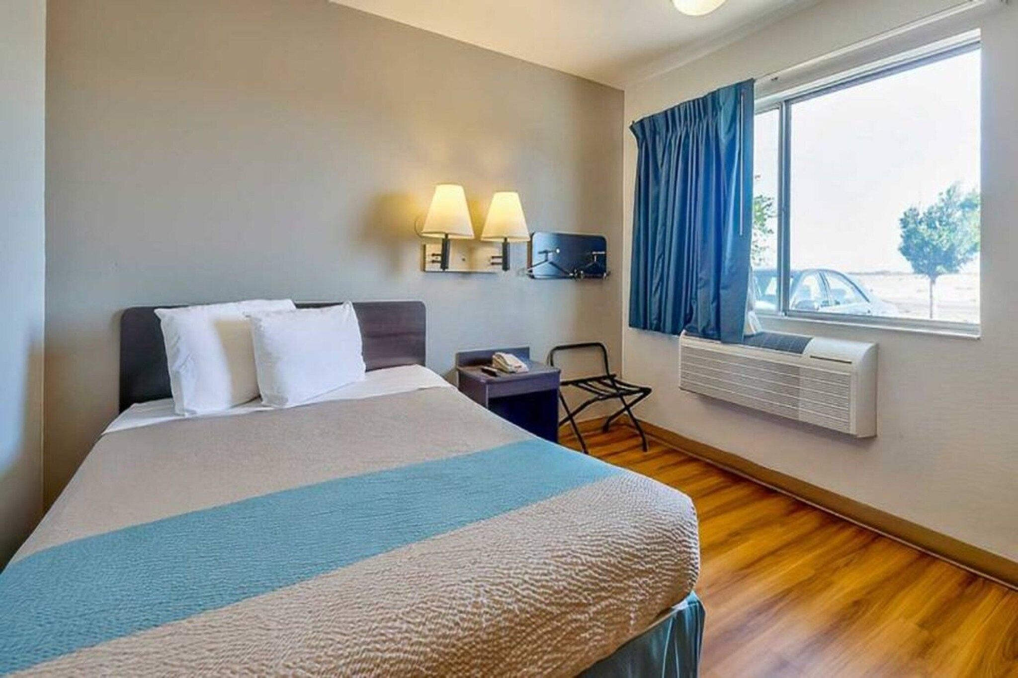 A hotel room with a bed, bedside lamps, a desk, a chair, a window with curtains, and an air conditioning unit.