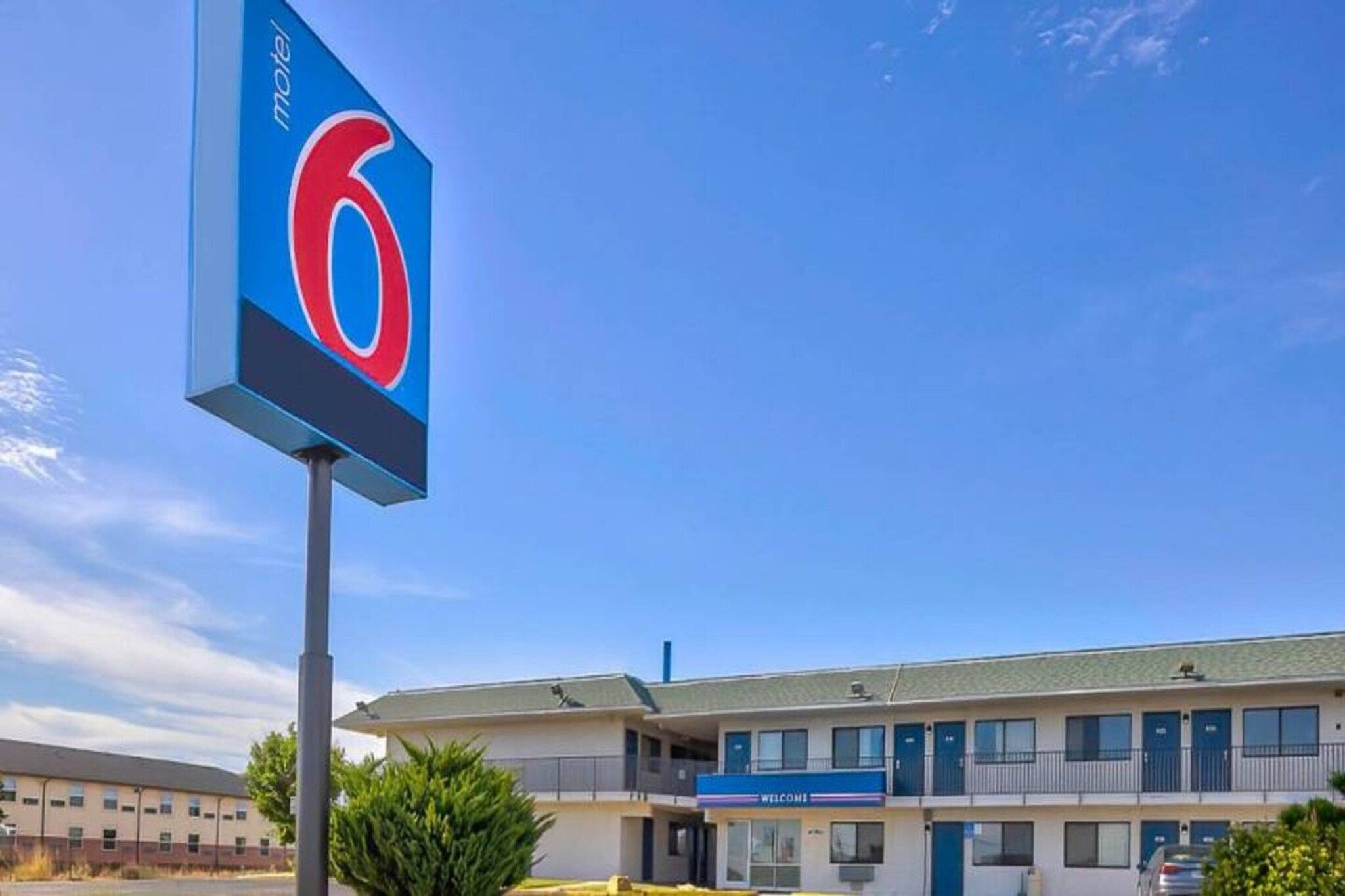 Um motel com um grande letreiro com o número 6, um estacionamento e um prédio com uma placa de 'Welcome'.