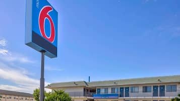 Um motel com um grande letreiro com o número 6, um estacionamento e um prédio com uma placa de 'Welcome'.