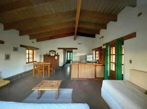 Ein Zimmer mit Holzbalkendecke, einem Bett, einer Couch, einem Esstisch mit Stühlen und einer Küchenzeile.
