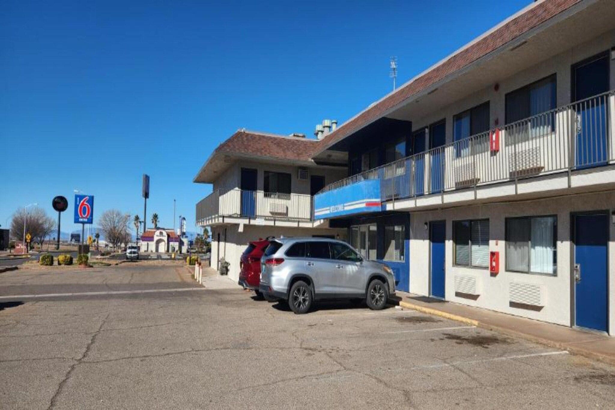 Un motel de dos pisos con estacionamiento, una camioneta SUV roja estacionada al frente y un cielo azul despejado.