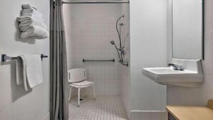 Een badkamer met een douche, een witte stoel, een wastafel en handdoeken aan haken.