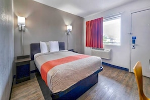 Quarto de hotel com cama, luminárias de cabeceira, janela com cortinas, aparelho de ar condicionado e uma cadeira.