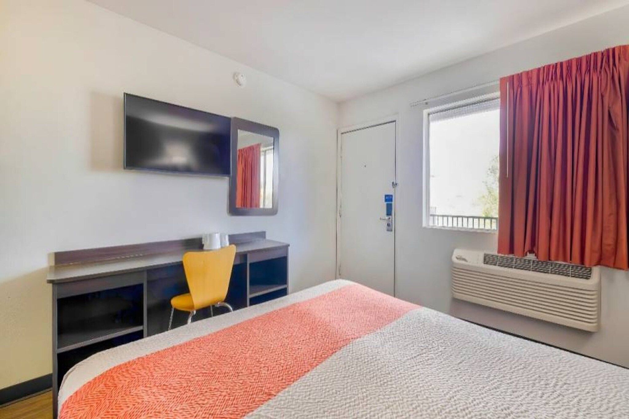Ett hotellrum med en säng, ett skrivbord, en stol, en spegel, ett fönster med gardiner och en luftkonditioneringsanläggning.