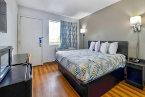 Quarto de hotel com cama, escrivaninha, televisão, janela com cortinas e mesa de cabeceira com abajur.