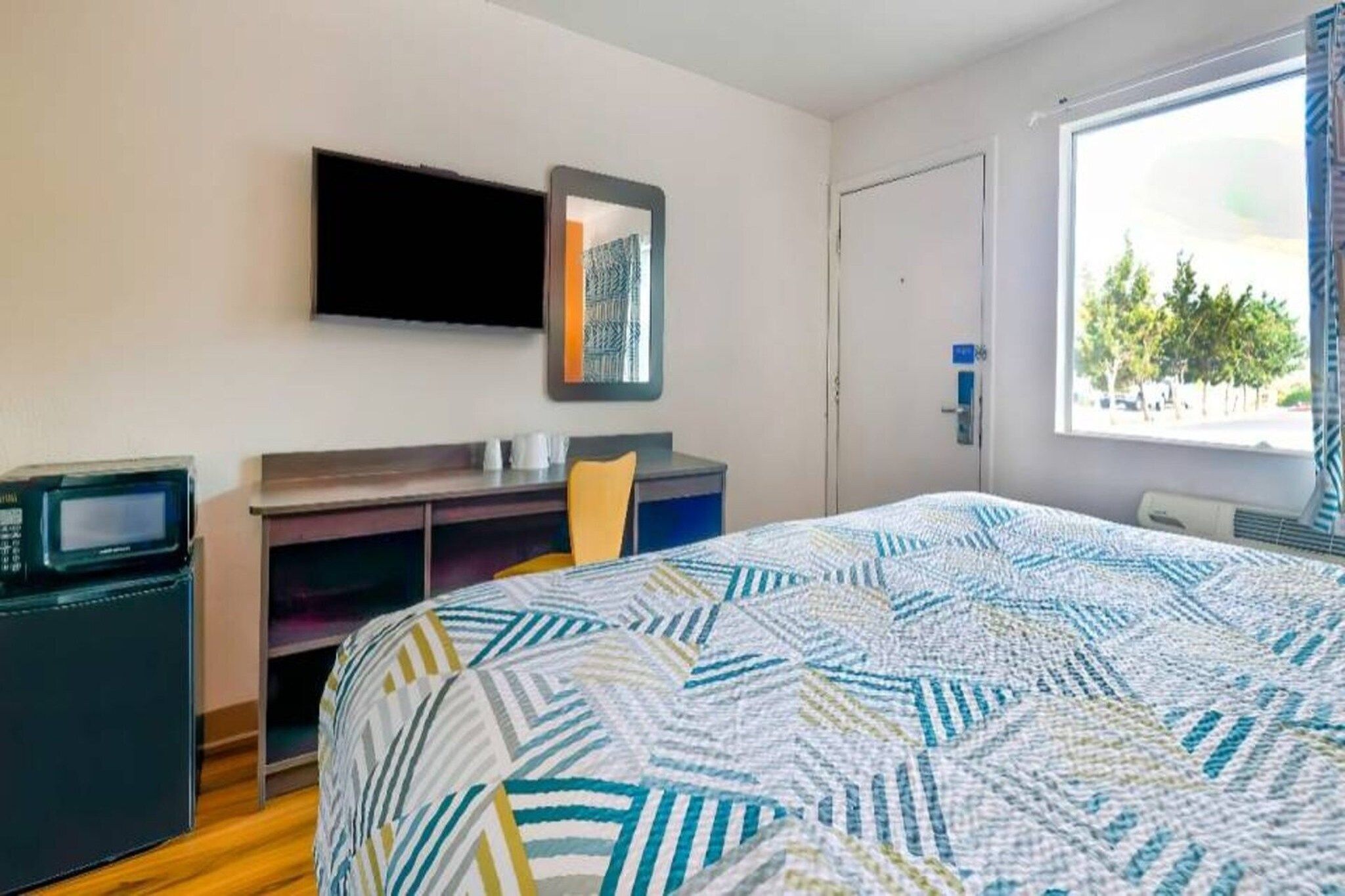 Habitación de hotel con cama, escritorio, silla, televisor y ventana con vista a árboles.