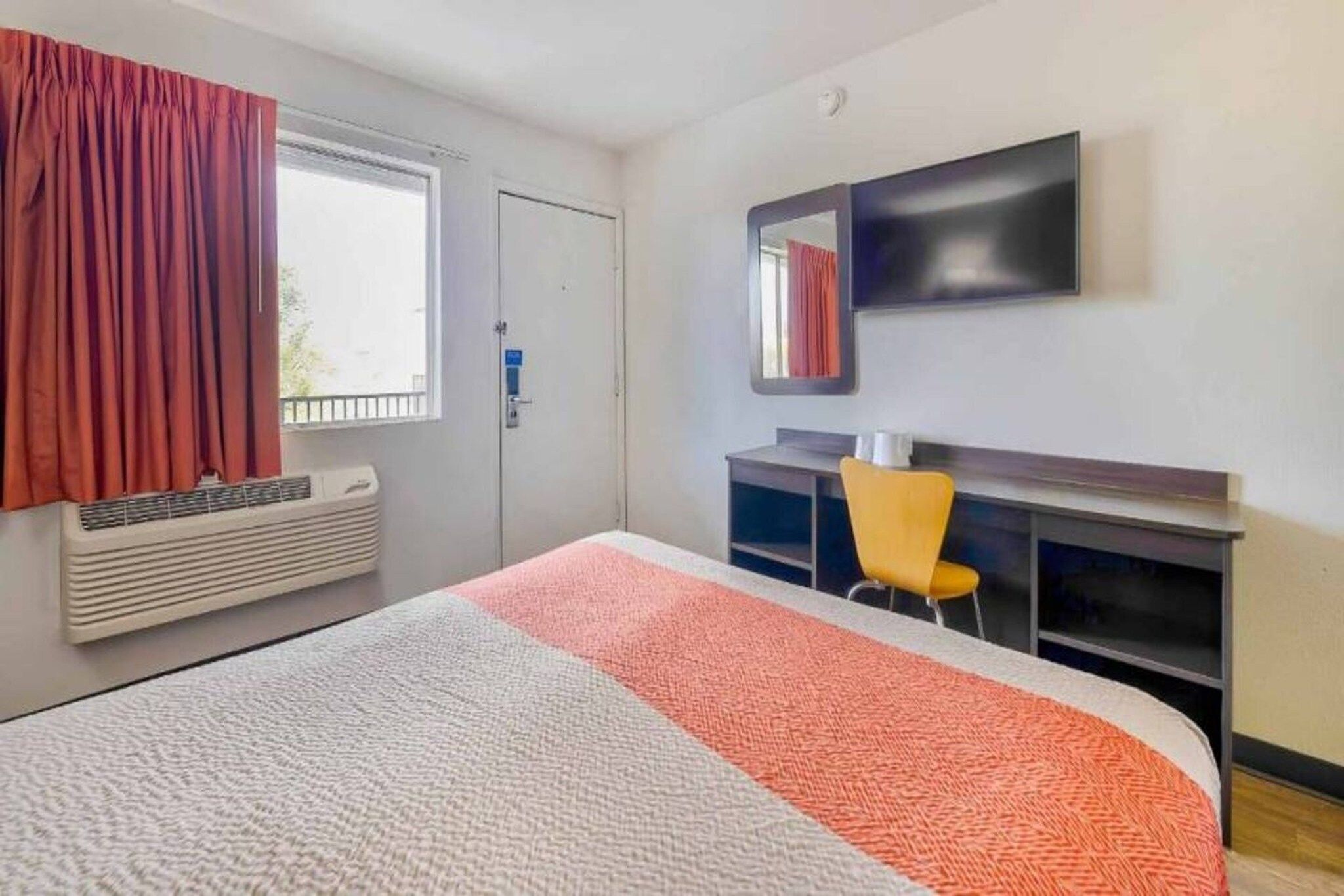 Ein Hotelzimmer mit Bett, Schreibtisch, Stuhl, einem Fenster mit Vorhängen, einem Heizkörper und einem an der Wand befestigten Fernseher.