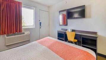 Ein Hotelzimmer mit Bett, Schreibtisch, Stuhl, einem Fenster mit Vorhängen, einem Heizkörper und einem an der Wand befestigten Fernseher.