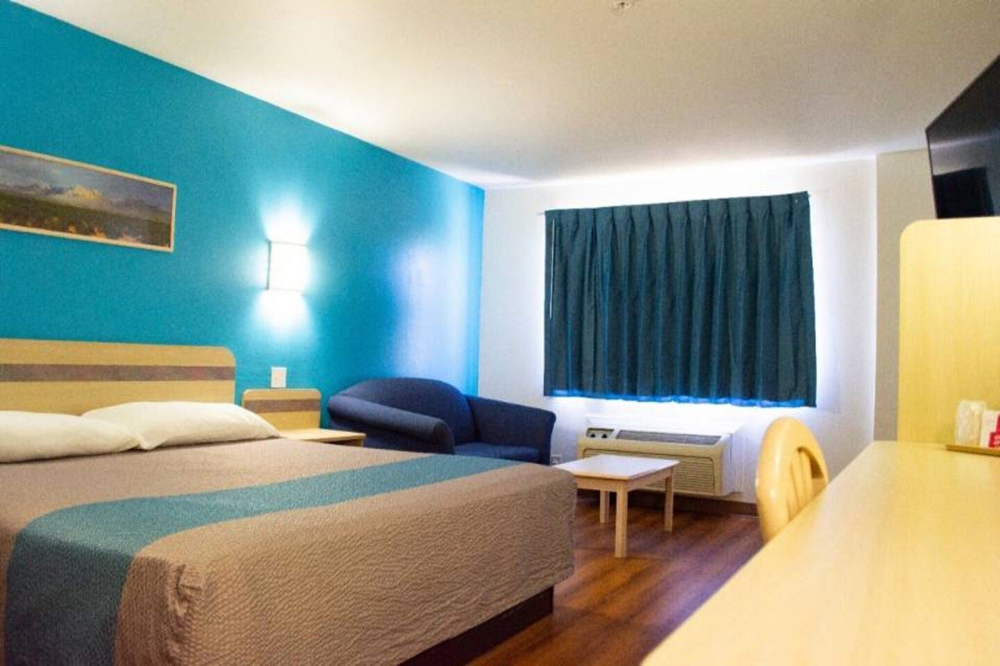 Habitación de hotel con una cama, un escritorio, una silla, una ventana con cortinas y una lámpara fijada a la pared.