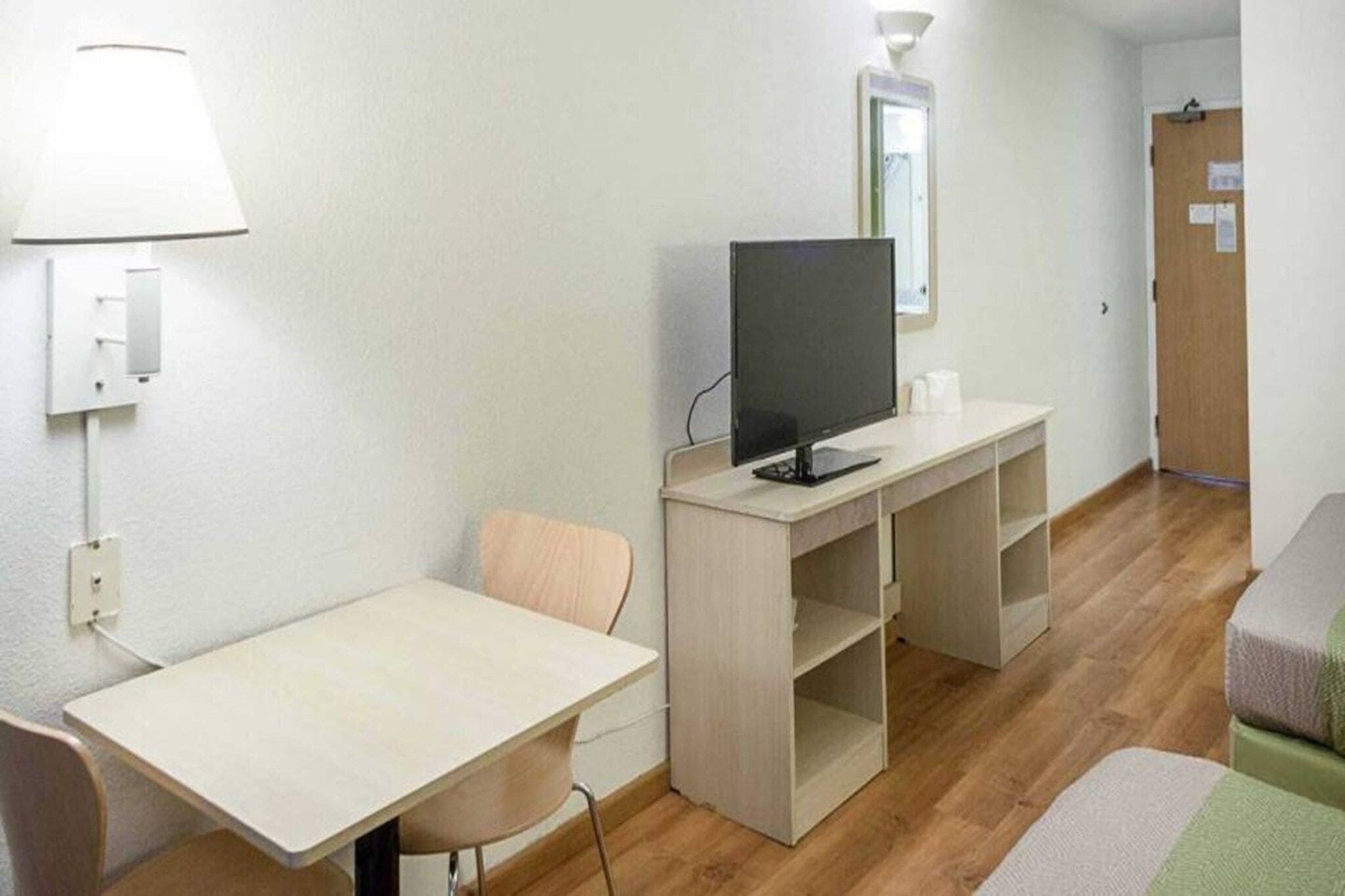 Una stanza piccola e modesta con una scrivania, una sedia, televisione e uno specchio appeso al muro.