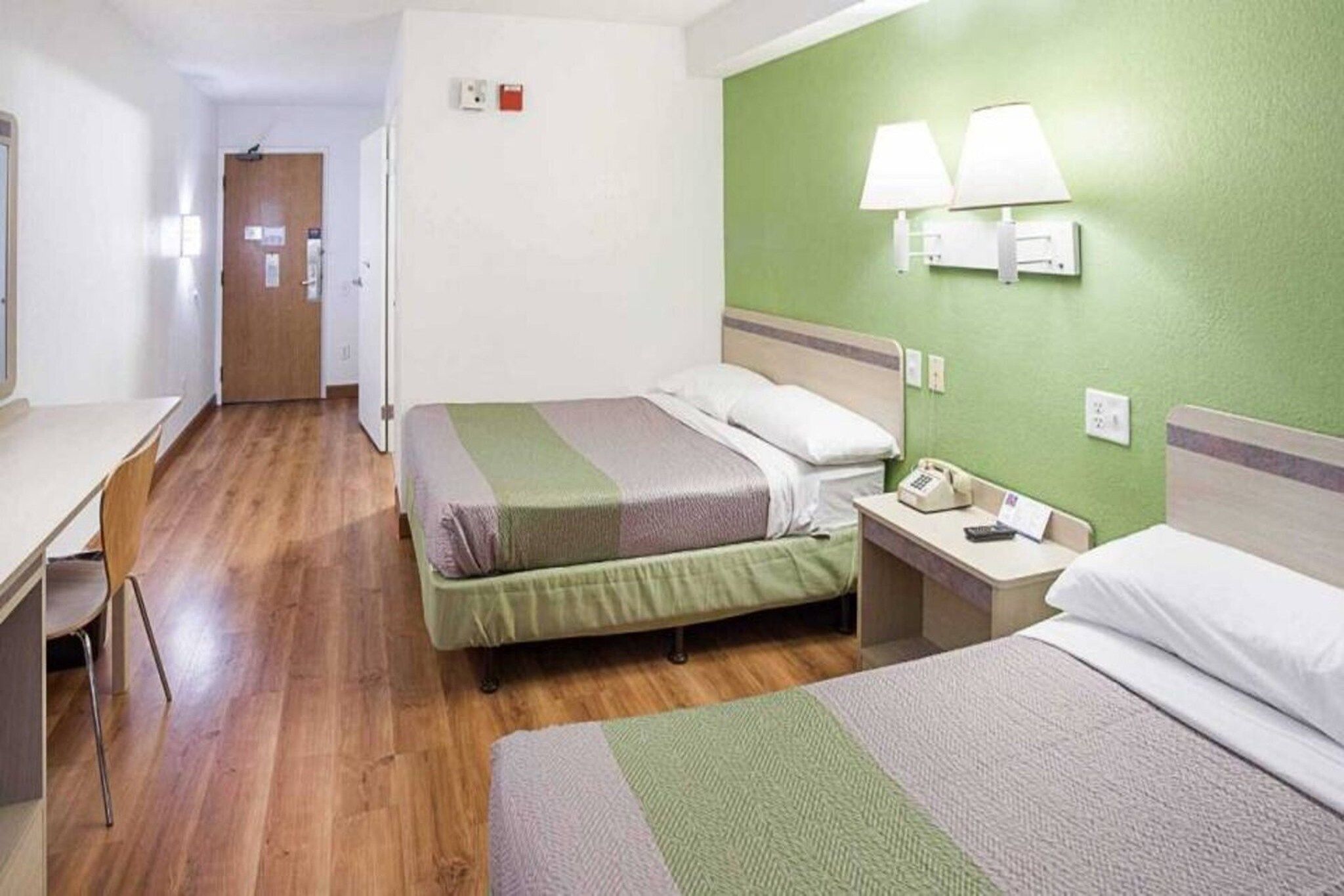 A hotel room with two beds, a desk, and a chair. There is a door leading to another room and a wall-mounted phone.